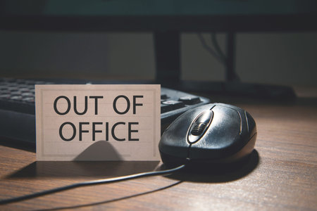Out of office sign on wooden desk with computer mouse and keyboard.