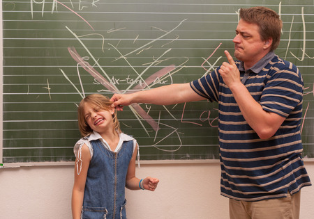 Angry teacher man pulling a naughty girls ear in front of the chalkboard.