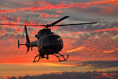 A black and red helicopter is silhouetted against a vibrant orange and red sunset sky.  The helicopter is in flight, and the image showcases its detailed structure.の素材