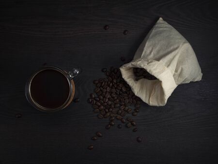 glass mug with fresh black natural coffee, a bag with coffee grains and sprinkled grains of roasted coffee on a wooden table.の写真素材