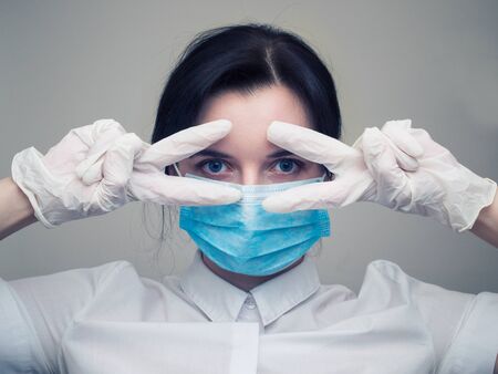 Young woman patient in a medical mask puts on protective surgical sterile gloves on her arm, on gray background, protection against coronovirusの写真素材