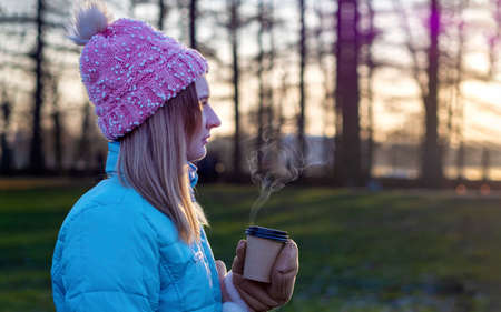 A beautiful cute girl in a hat and a down jacket holds a paper cup with hot coffee or tea. Frosty sunny morning Winter season.の写真素材