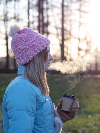 A beautiful cute girl in a hat and a down jacket holds a paper cup with hot coffee or tea. Frosty sunny morning Winter season.の写真素材