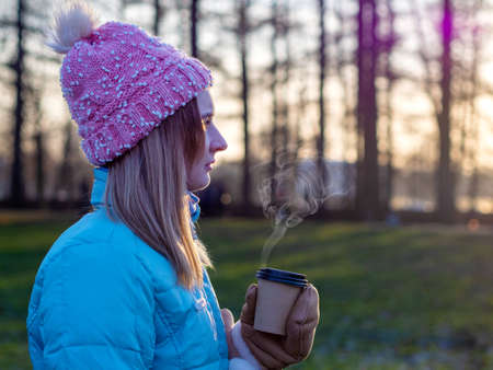 A beautiful cute girl in a hat and a down jacket holds a paper cup with hot coffee or tea. Frosty sunny morning Winter season.の写真素材