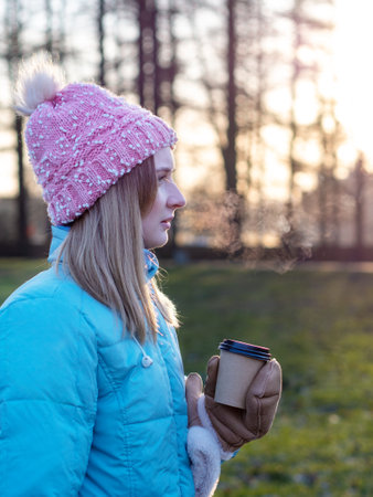 A beautiful cute girl in a hat and a down jacket holds a paper cup with hot coffee or tea. Frosty sunny morning Winter season.の写真素材