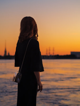 A young elegant woman stands on the embankment and looks at the orange sunset on the Neva River and the Peter and Paul Fortress. St. petersburgの写真素材