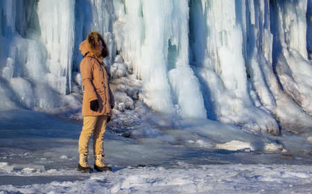 A young traveler woman in a jacket with a hood and fur stands at a frozen waterfall surrounded by iceの写真素材