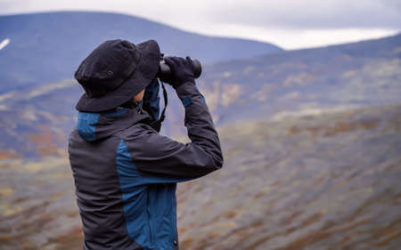 A woman in a hat on a mountain slope looks through binoculars in autumn against the backdrop of a lake and mountains. view from the back.の写真素材