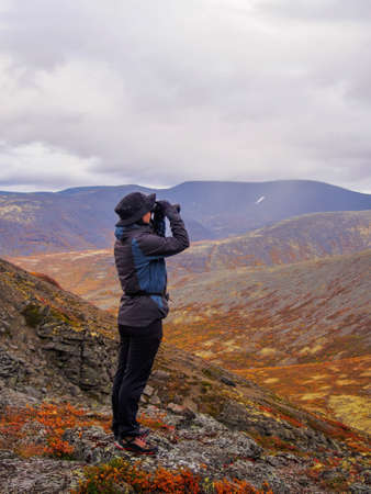 A woman in a hat on a mountain slope looks through binoculars in autumn against the backdrop of a lake and mountains. view from the back.の写真素材