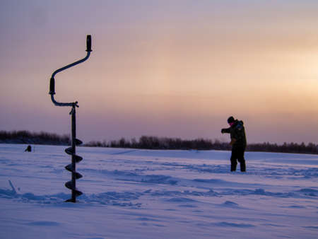 Ice drill on a frozen lake with a fisherman in the background.の写真素材