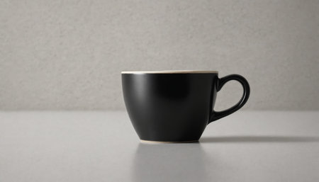 A black mug sits on a white table, illuminated by a single beam of light. The mug casts a long shadow on the table, creating a stark contrast between light and dark.の素材