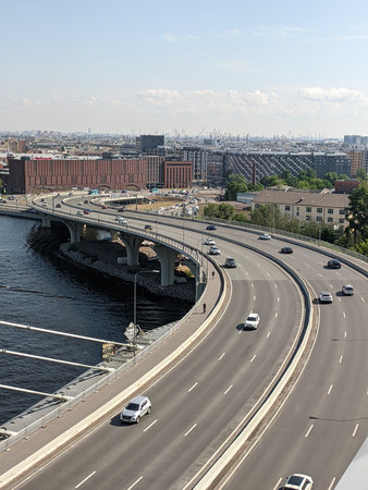 Bright sunlight illuminates a bustling city as cars travel along a curved highway beside a calm...