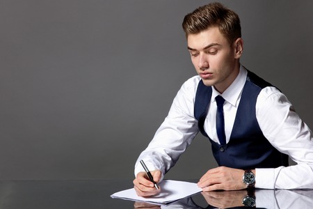 A model of a man sits at a table in a suit, holds a pen, on the table a white sheet of paperの写真素材