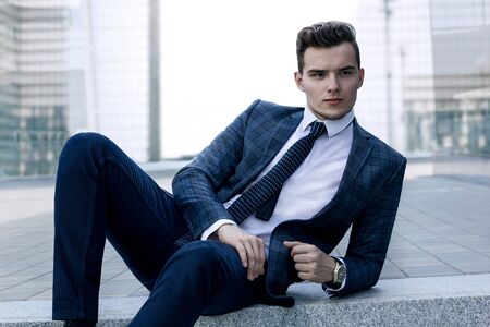 fashionably dressed man in a checkered jacket with a tie and a wrist watch posing while sitting against the backdrop of the cityの写真素材