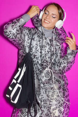 Girl listens to music on a pink and purple background and dances in a fashionable raincoat and headphonesの写真素材
