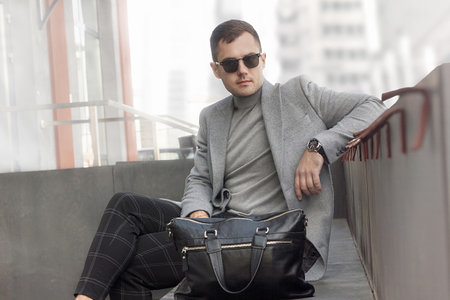 A man in a gray coat and sunglasses sits in the city. Demi-season fashion.の写真素材
