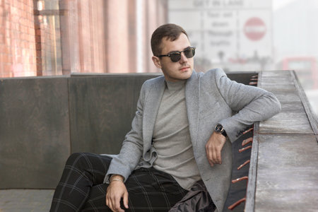 A man in a gray coat and sunglasses sits in the city. Demi-season fashion.の写真素材