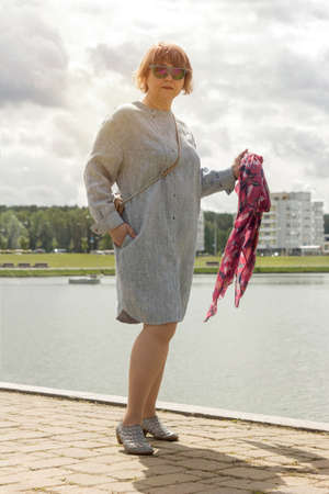 Mature plus size woman walks along the shore of the lake in the park in a trendy dress in summer. Urban summer fashion.の写真素材