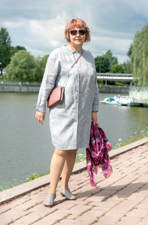Mature plus size woman walks along the shore of the lake in the park in a trendy dress in summer. Urban summer fashion.の写真素材