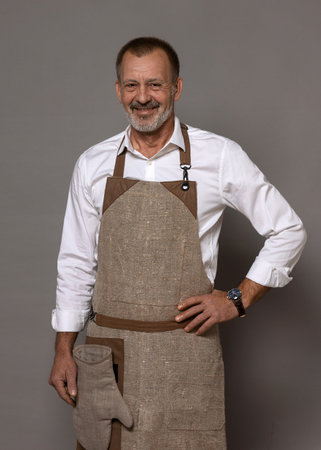 A mature man in a chef's apron with an oven mitt is standing and smiling. isolated background.の写真素材