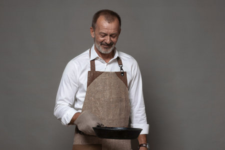 A mature man in a chef's apron holds a cast-iron frying pan with an oven mitt. isolated background.の写真素材