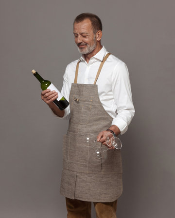 Respectable man in a linen apron holds wine glasses and a bottle of wine. Sommelier or waiter in joy and hospitality.の写真素材