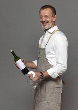 Respectable man in a linen apron holds wine glasses and a bottle of wine. Sommelier or waiter in joy and hospitality.の写真素材