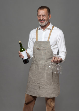 Respectable man in a linen apron holds wine glasses and a bottle of wine. Sommelier or waiter in joy and hospitality.の写真素材