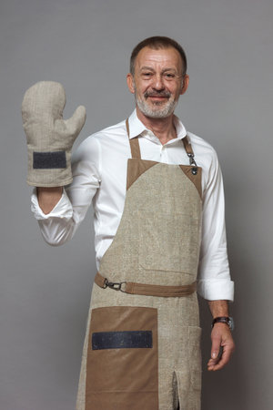 Mature man in a chef's apron waves, greets with an oven mitt and smiles. Three quarter portrait.の写真素材