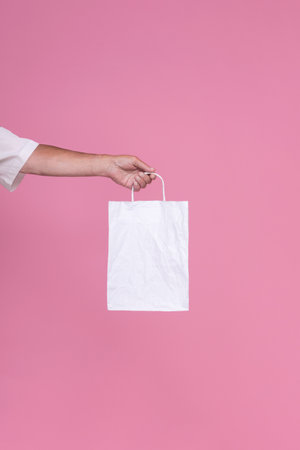 Hand holding empty white paper bag for advertising and branding mockup template isolated on pink...