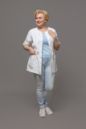 Friendly female doctor posing from a standing position, woman in uniform, full length, gray studio background banner.の写真素材