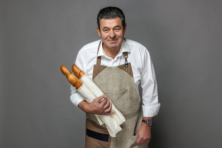 Bread seller, baguette in bakery on gray background. Mature French baker holding baguette in hands.の写真素材