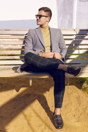 Man in coat and sunglasses sitting on bench in city. Demi-season fashion, spring, autumn.の写真素材
