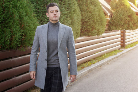 Fashionable man in a gray coat walks against the background of a thuja alley. Advertising, luxury...