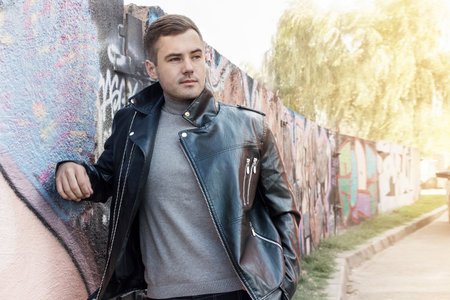 Fashionable man in a black leather kosovorotka against the background of a city wall with graffiti. Advertising, luxury life.の写真素材