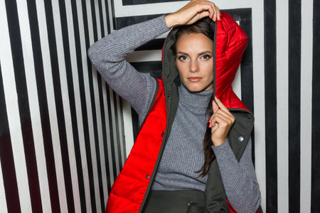 Young woman in red quilted down vest poses gracefully on creative striped background. Sitting, with beautiful makeup.の写真素材