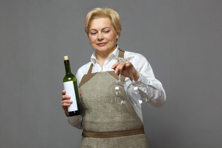 Elegant mature waitress in apron and white shirt holding bottle of wine and glasses. Isolated image. Holiday, service.の写真素材