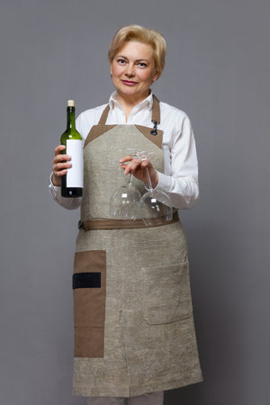 Elegant mature waitress in apron and white shirt holding bottle of wine and glasses. Isolated image. Holiday, service.の写真素材