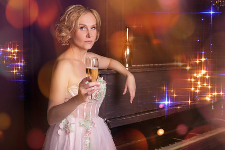 Woman in pink dress leaning on piano with glass of white wine in festive atmosphere of lights. Advertisement of music salon or evening.の写真素材
