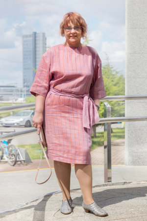 A smiling woman wearing a pink dress and glasses stands outdoors on a sunny day. In the summer, a mature plus size woman stands on the sidewalk among office buildings in a pink dress. Urban summer fashion.の写真素材