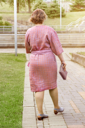 A woman in a pink dress is walking away on a paved path in a park-like setting. In the summer, a mature plus size woman walks in a city park in a pink dress. Urban summer fashion.の写真素材