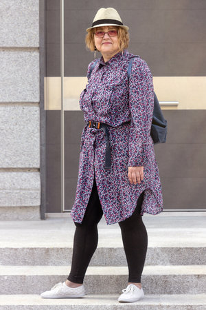 Stylish plus-size woman stands on the street in an small urban setting in the city, wearing a long shirt with a floral print and a white hat.の写真素材