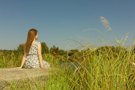 A girl with long hair sits relaxed on the river bank on a hot summer day. View from behind.の写真素材
