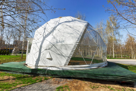 A white geodesic dome located outdoors under a clear blue sky. The shadow of the tree is cast on the...