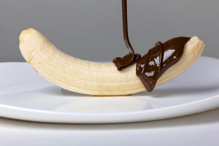 A close-up of a banana covered with an even layer of melted chocolate, dripping down it onto a clean white plate. The series of technological processes consists of 9 photos, the third photo.の写真素材
