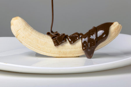 A close-up of a banana covered with an even layer of melted chocolate, dripping down it onto a clean white plate. The series of technological processes consists of 9 photos, the fourth photo.の写真素材