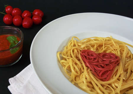 Red linguine heart-shaped with yellow linguine arranged on a white plate, horizontal, partial shot, black backgroundの写真素材