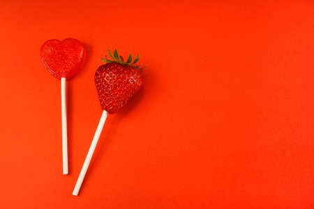 2 lollipops, a fresh strawberry on a stick and a heart-shaped lollipop, horizontal, red background, still free spaceの写真素材