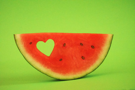 Piece of watermelon against a green background, horizontal, with a heart-shaped punched holeの写真素材