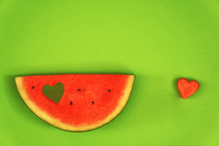 Piece of watermelon against a green background, horizontal, with a heart-shaped punched hole and with a punched-out melon heart next to itの写真素材